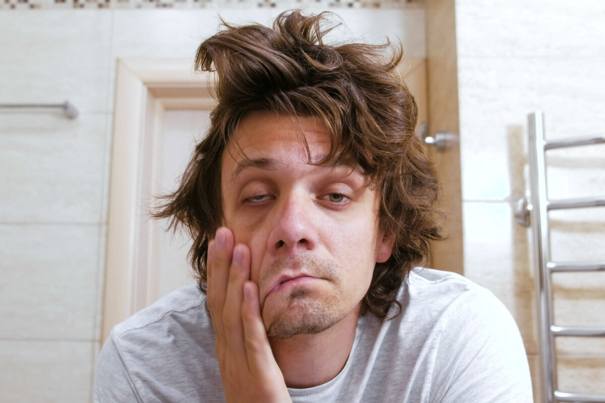 Sleepy shaggy young man looks at the mirror in bathroom in the morning